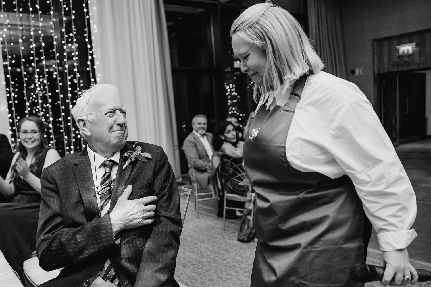 Black and white photo of a restaurant server having an animated conversation with an elderly guest at a dinner event. Wedding PHOTOGRAPHER D