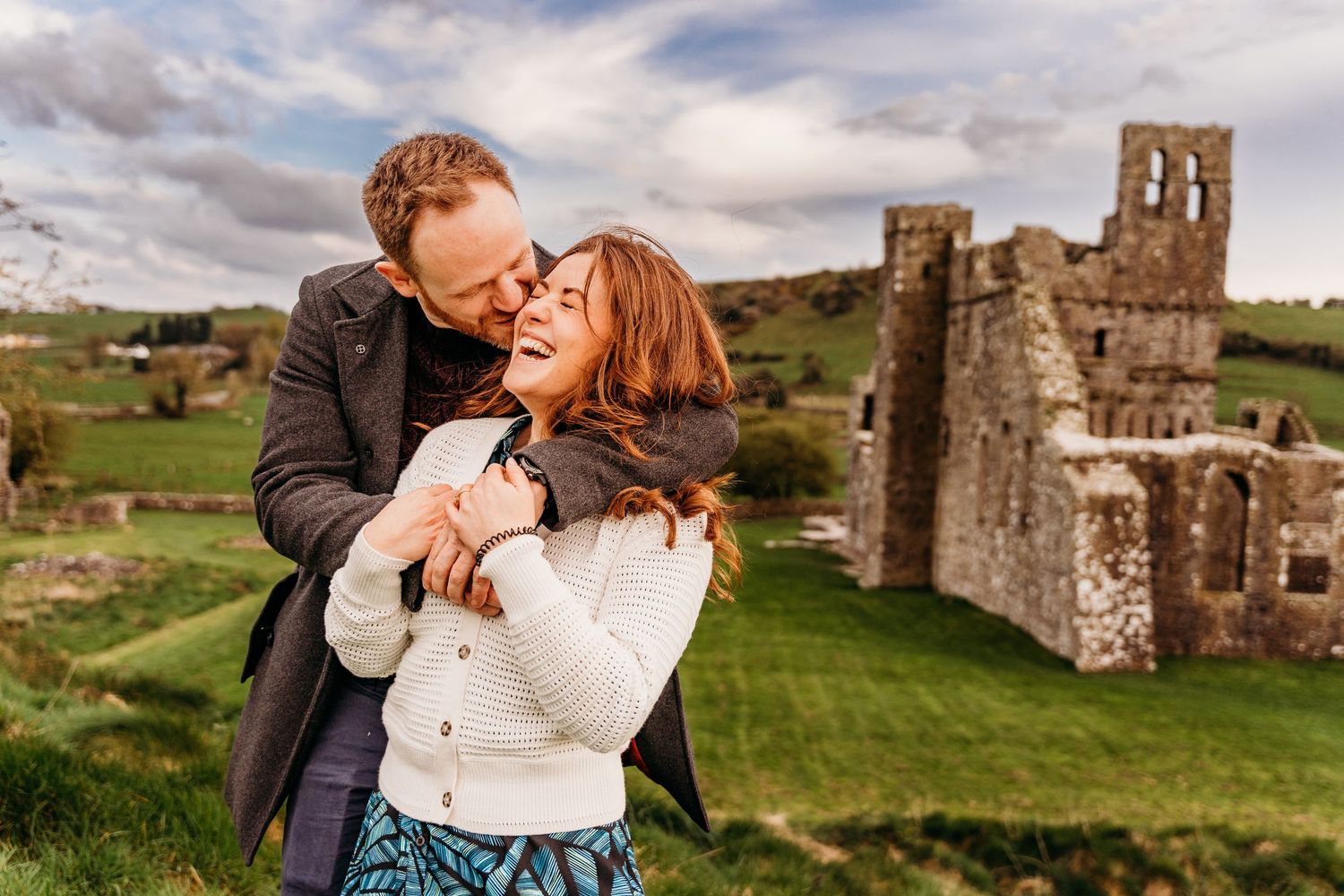 A couple shares an intimate moment in front of historic castle ruins on a green hillside under cloudy skies. PHOTOGRAPHER Dublin ireland