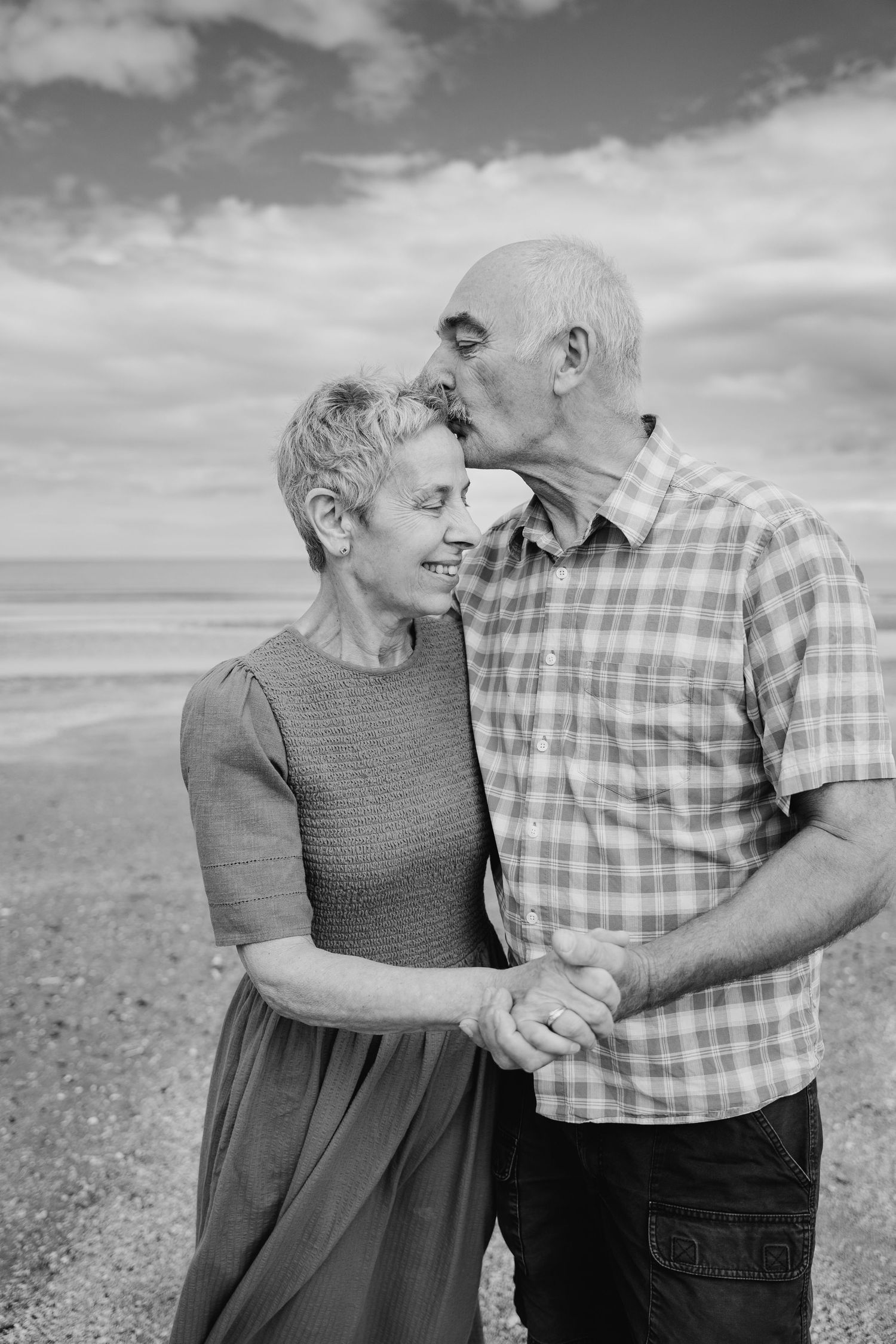 Black and white photo of a senior couple embracing on a beach, sharing an affectionate moment against a cloudy sky. PHOTOGRAPHER Dublin irel