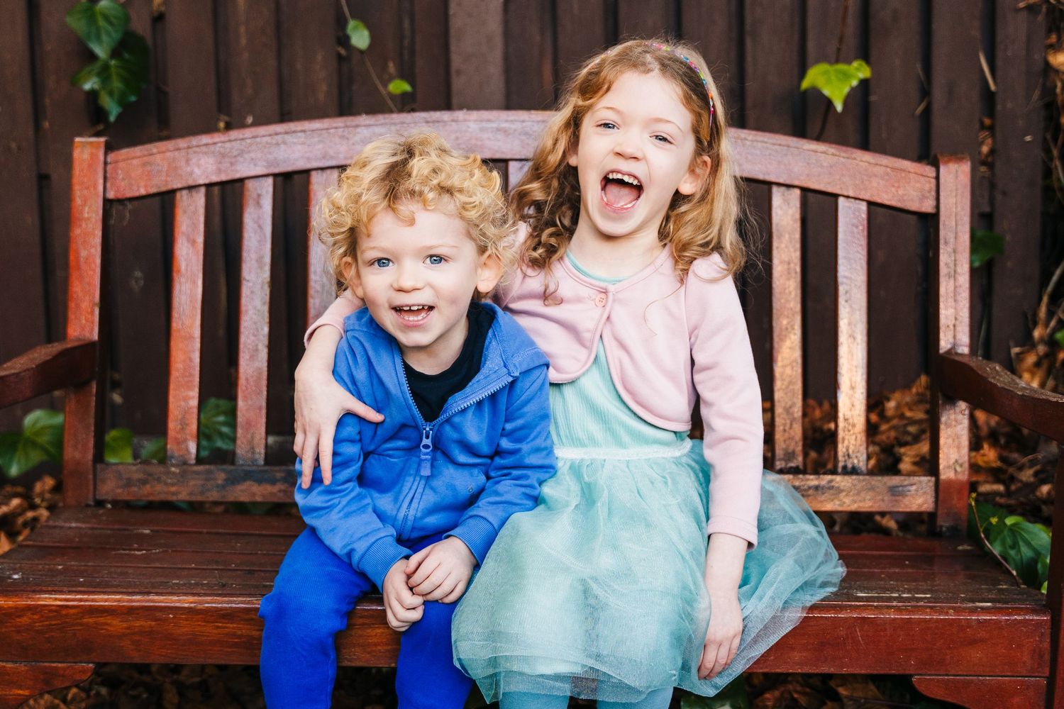Two children laughing joyfully while sitting on a wooden bench in a garden setting. Family PHOTOGRAPHER Dublin family PHOTOGRAPHER Dublin