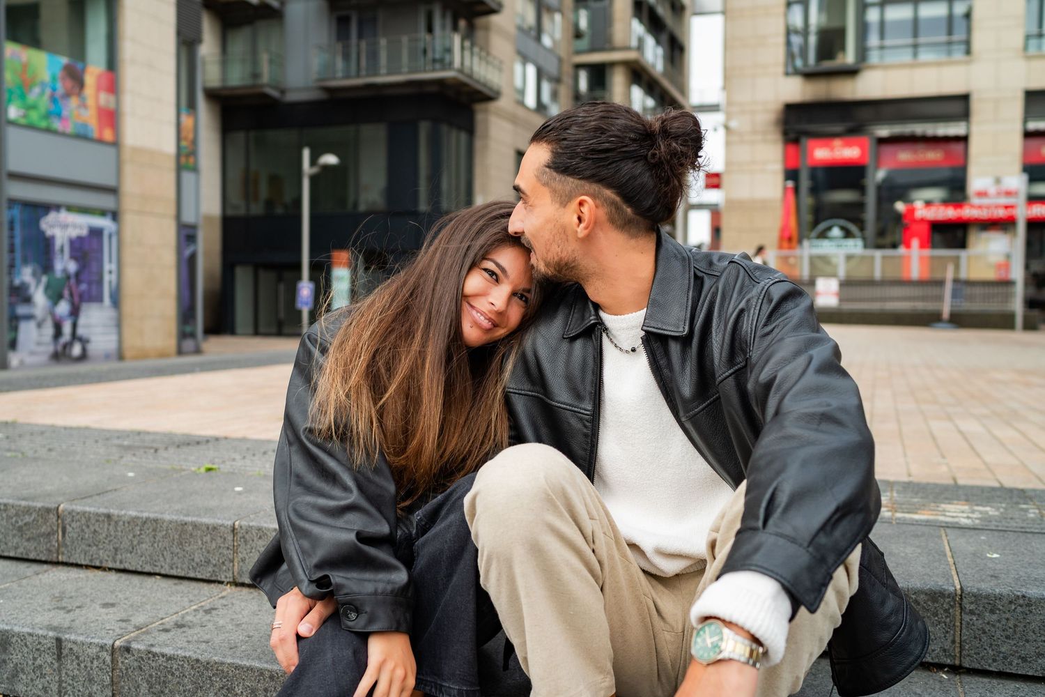 Couple sharing a tender moment outdoors on urban steps with city buildings in background.