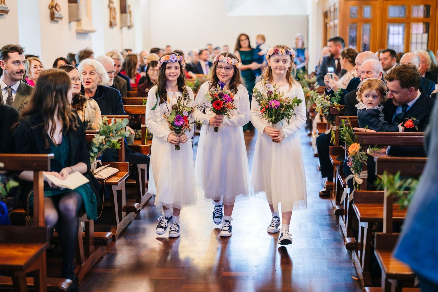 Bridesmaids in light dresses and sneakers walk down the church aisle holding colorful bouquets.
