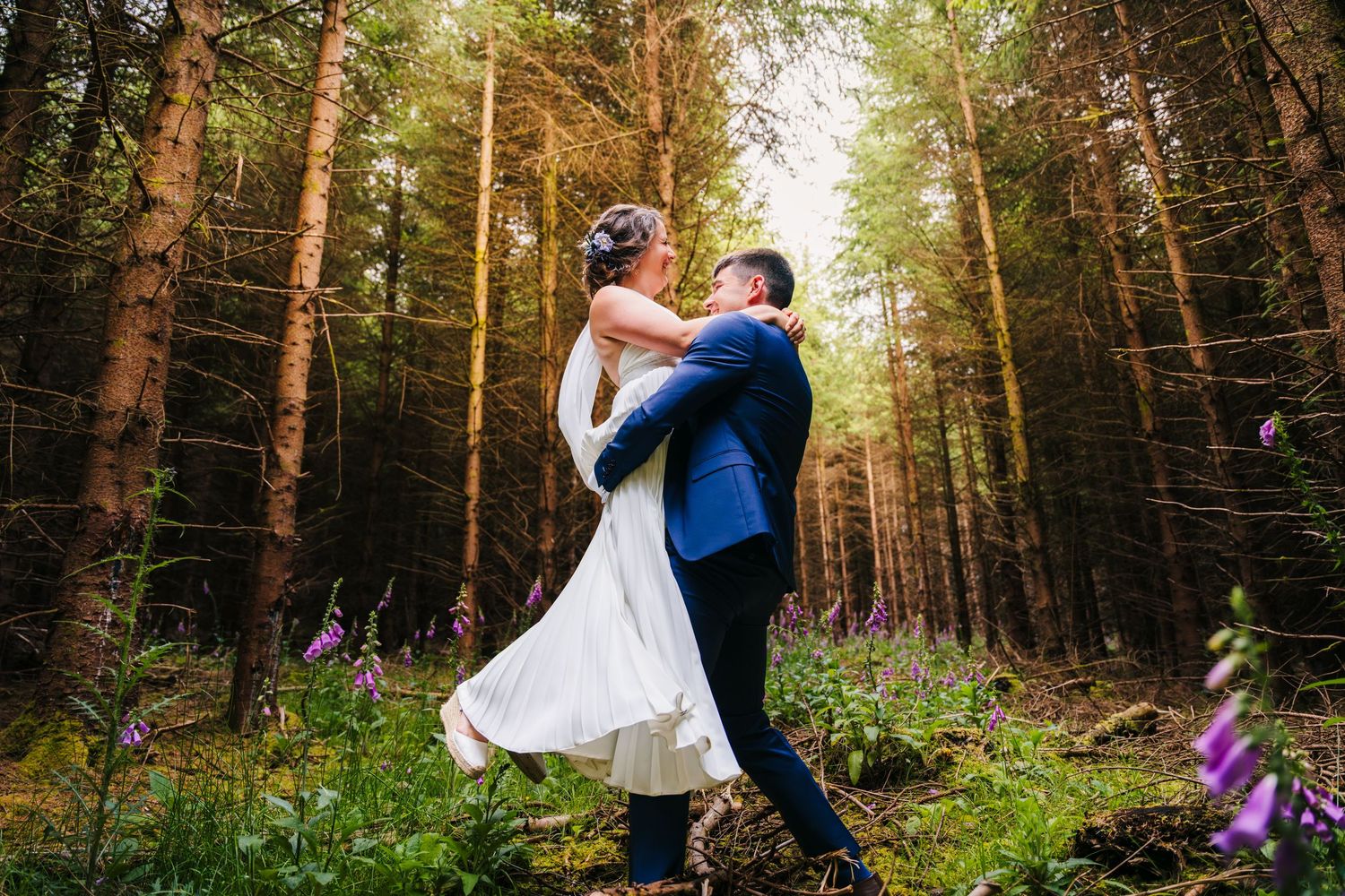 A couple in wedding attire share a romantic dance in a sunlit forest clearing surrounded by tall pine trees.