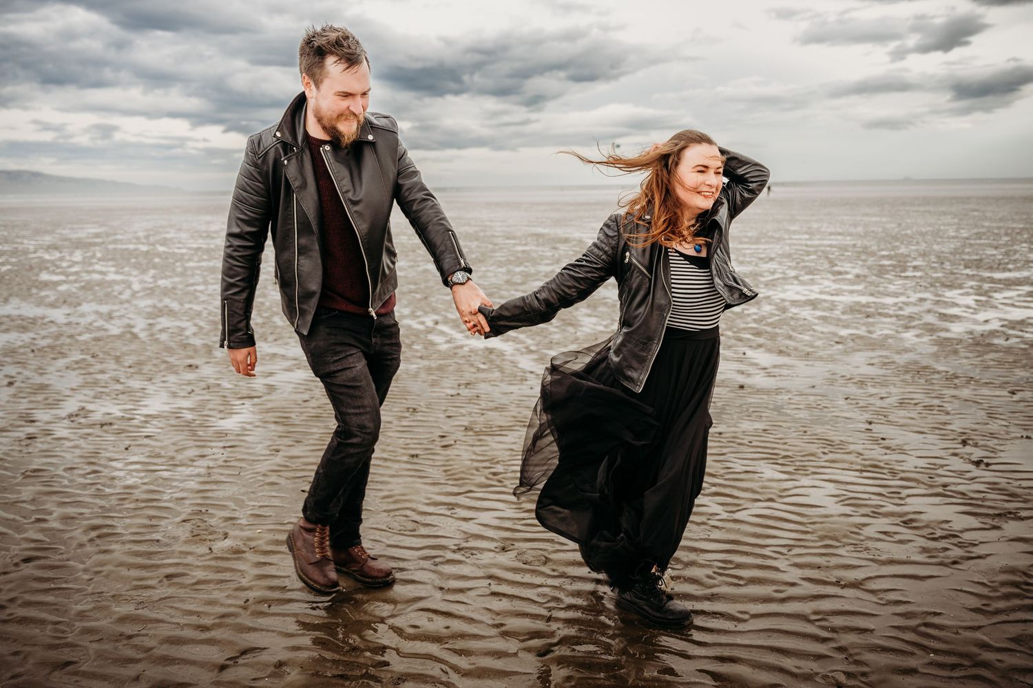 Two people playfully dance and twirl together on a moody beach under cloudy skies at sunset. engagement PHOTOGRAPHER Dublin