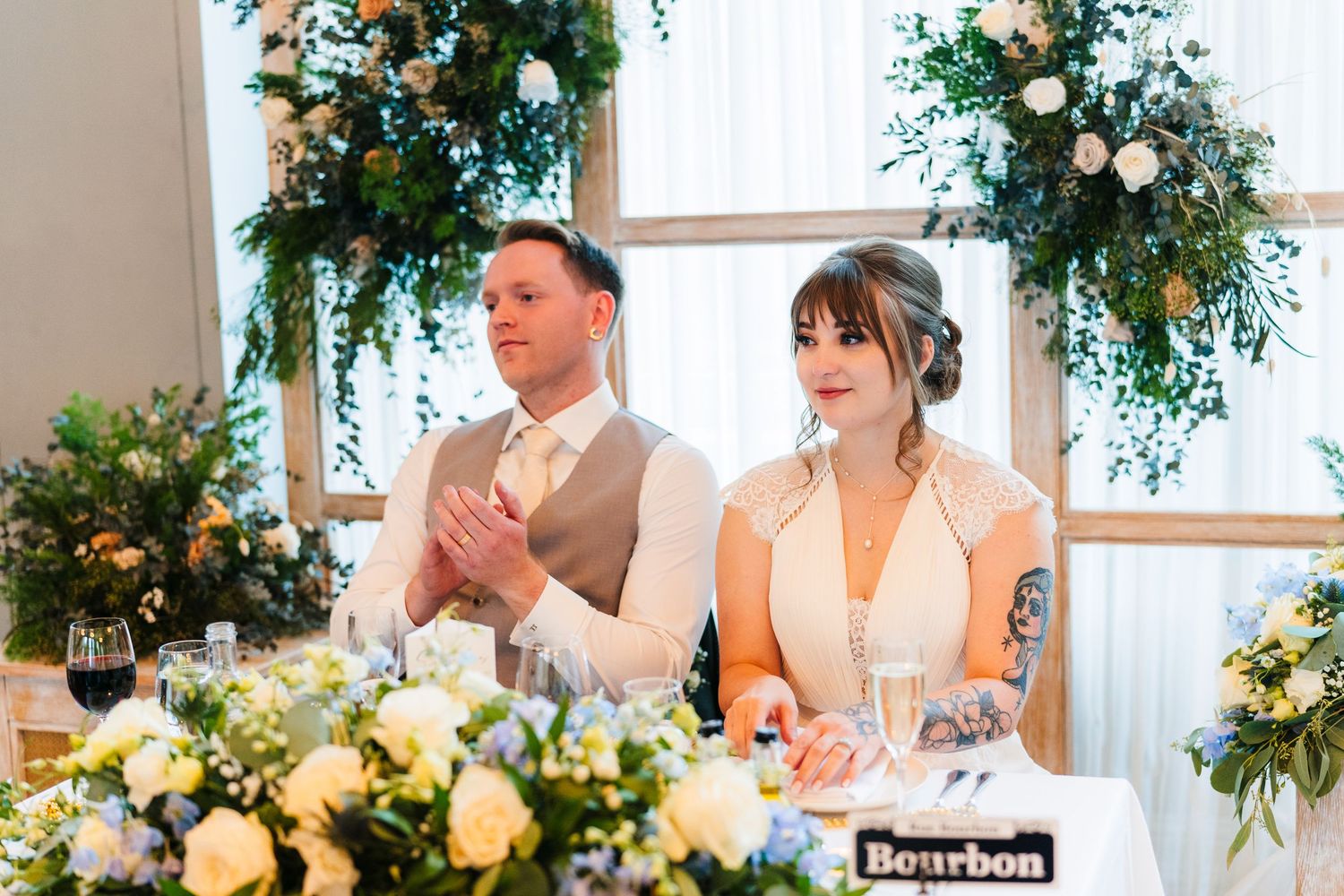 A couple seated at their wedding reception table under elegant floral arrangements clap during speeches.PHOTOGRAPHER Dublin Ireland Wedding