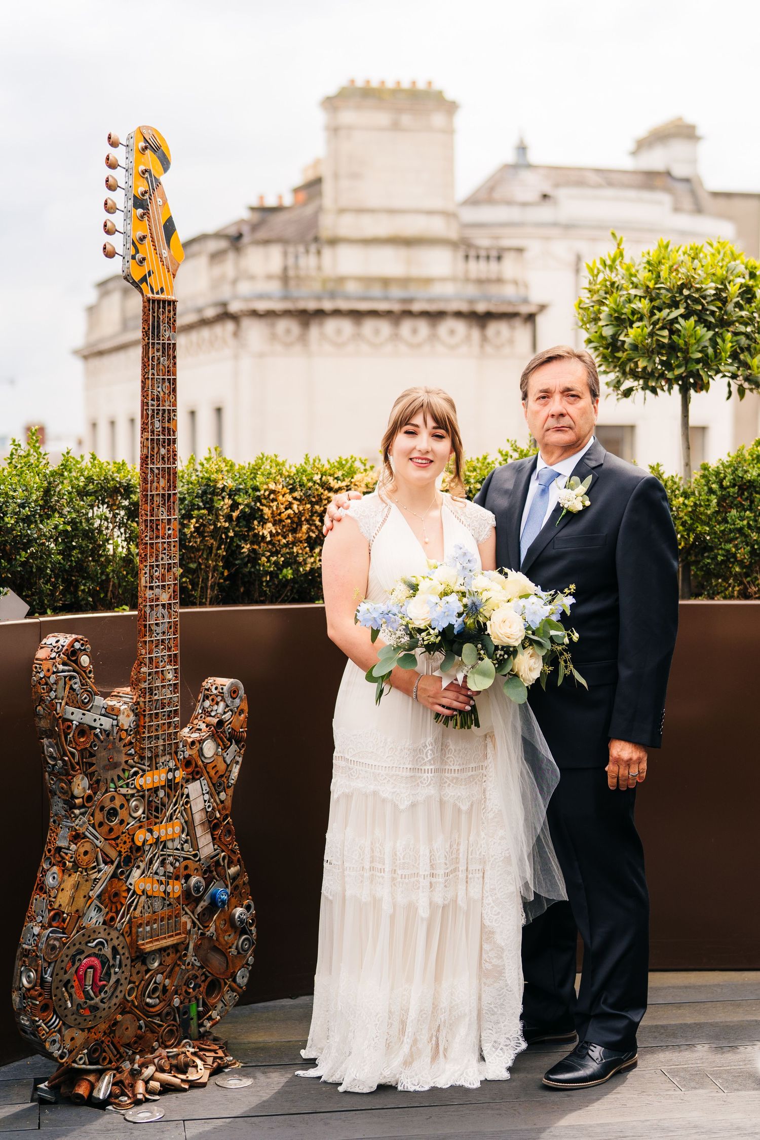 A decorative guitar stands next to a couple on their wedding day with a grand building in the background. PHOTOGRAPHER Dublin Ireland Weddin