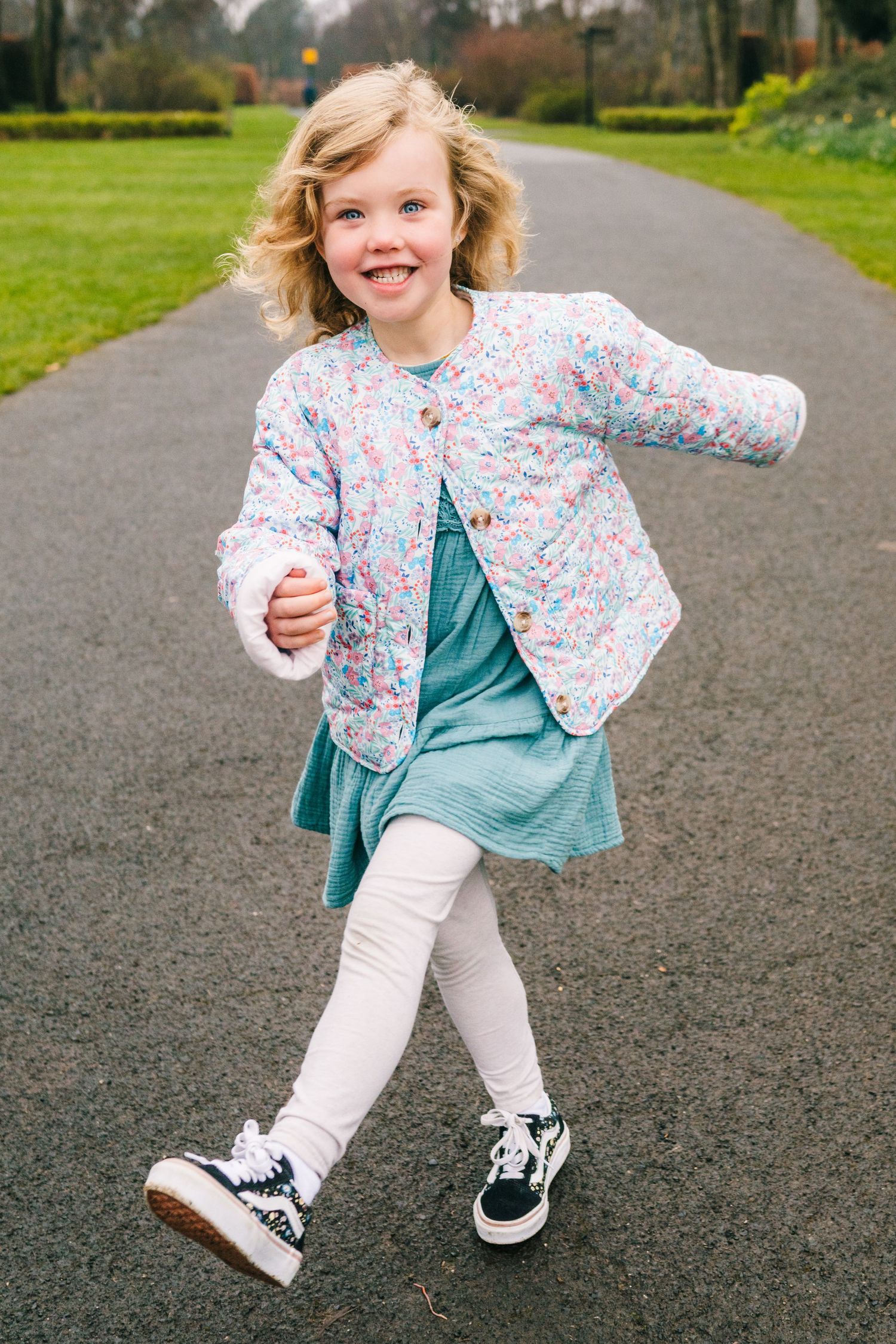 Family photographer Dublin Ireland  A playful child wearing a floral jacket and teal dress skips along a park path.