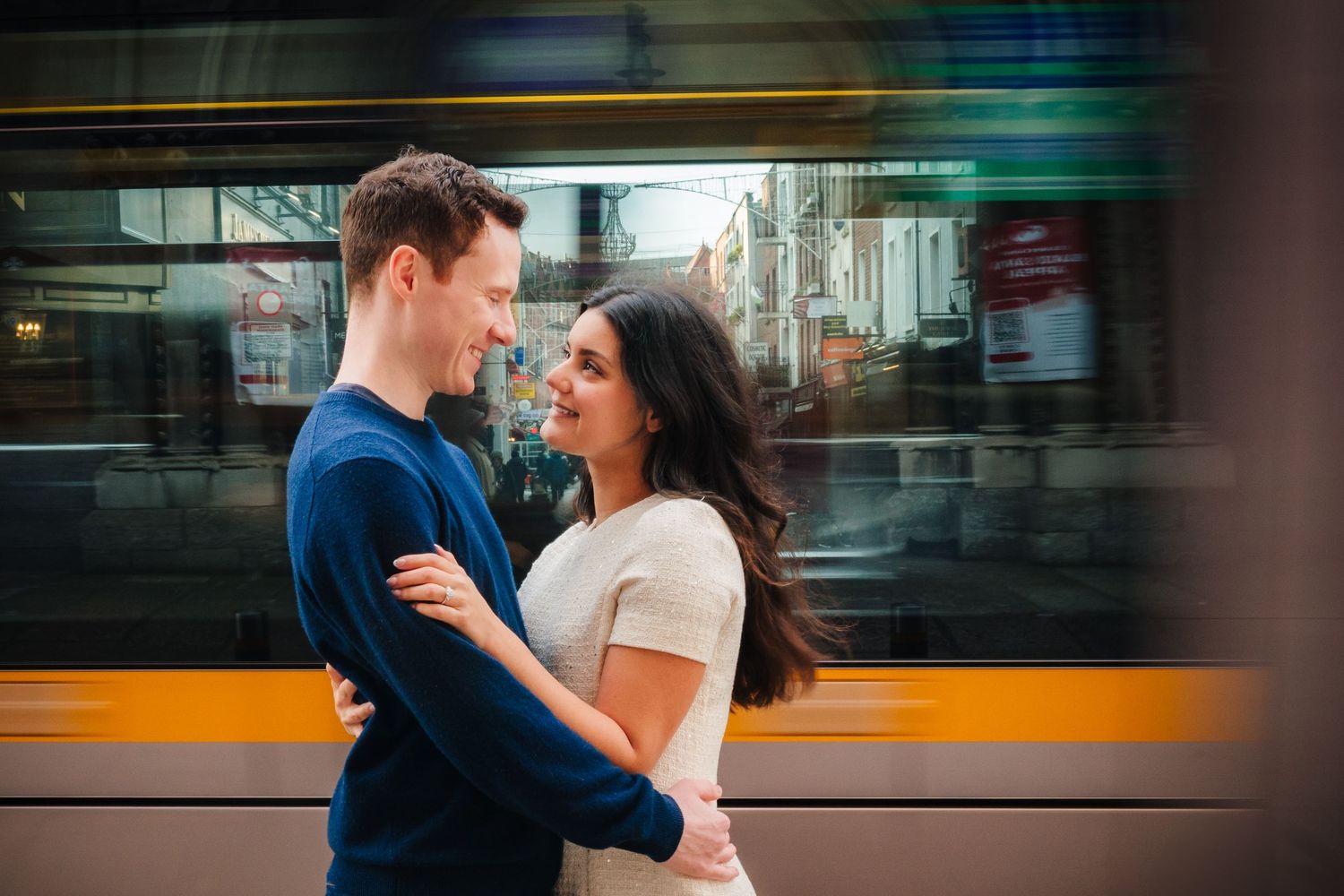 A couple embraces in front of a passing vibrant orange train in an urban setting. Surprise proposal dublin photography