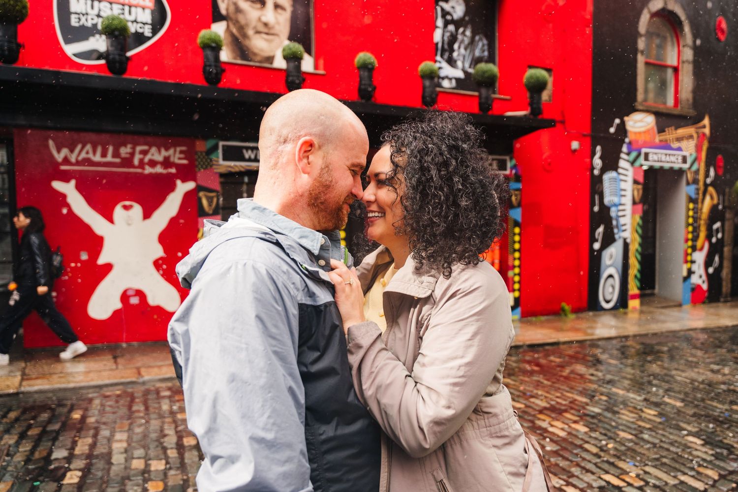 A couple shares a romantic moment in front of a vibrant red wall with umbrellas and street art in the background. Proposal Dublin Photograph