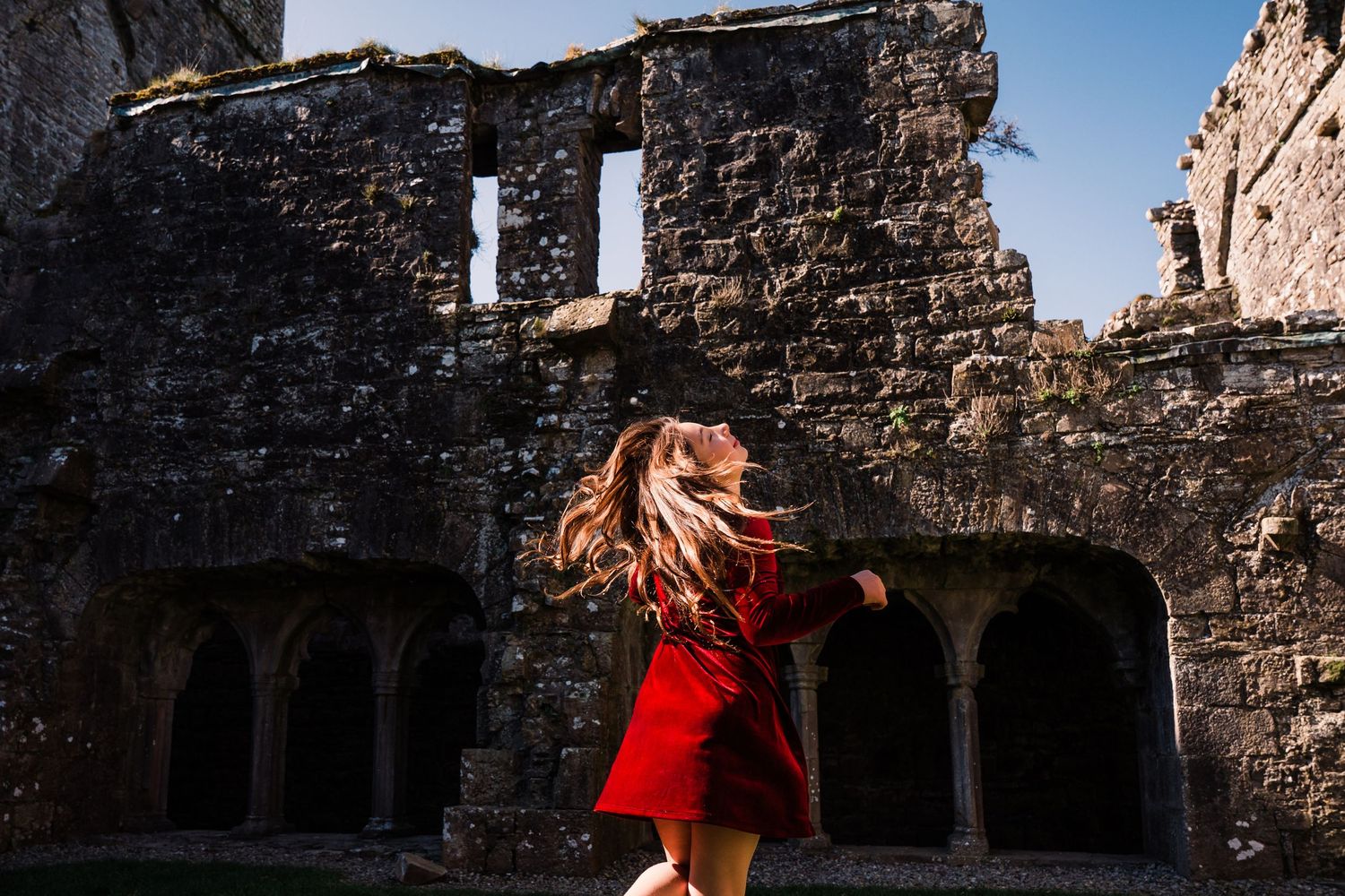 Person in flowing red dress twirling among ancient castle ruins against blue sky. Family PHOTOGRAPHER Dublin