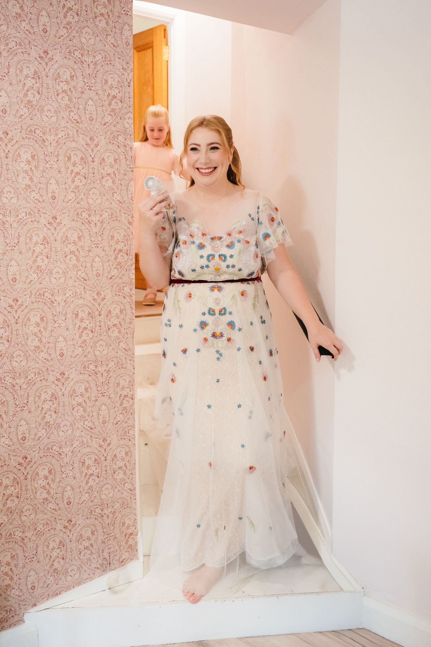 A white wedding dress with floral embellishments and tulle skirt displayed in a room with pink floral wallpaper. . Village Barn Tyrrellspass