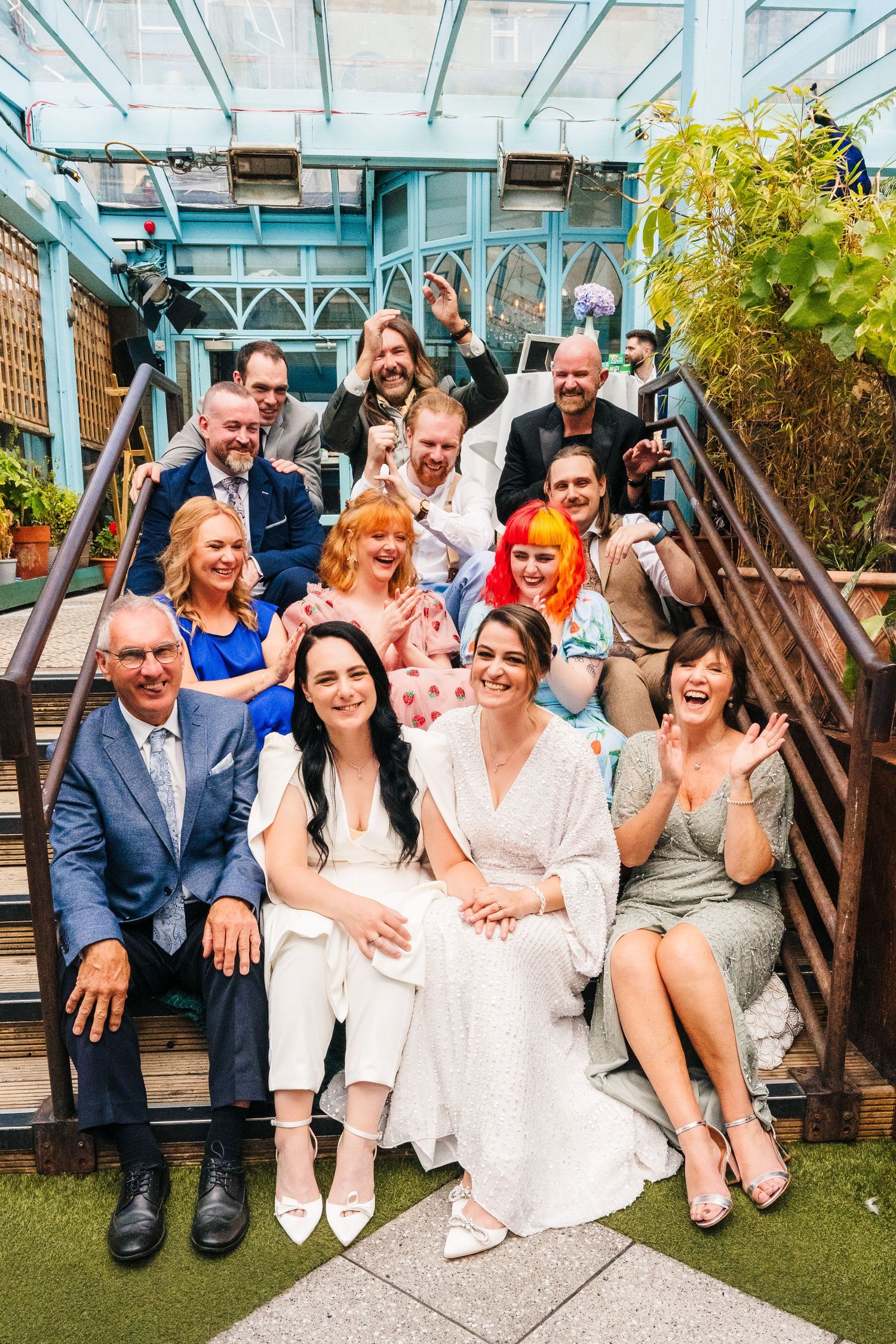 A joyful wedding group sitting together in a greenhouse setting with natural light. House Dublin Wedding Photographer