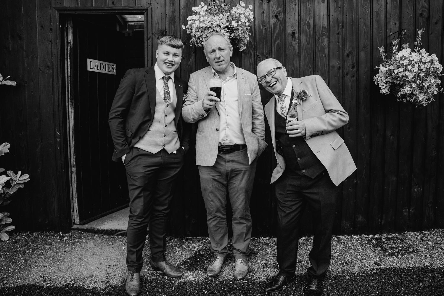 Three wedding guests in suits pose with drinks against a wooden wall at the reception. Wedding photographer Dublin