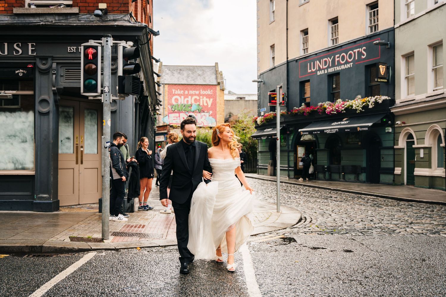 A couple walks across a cobblestone street in front of historic storefronts on their wedding day.