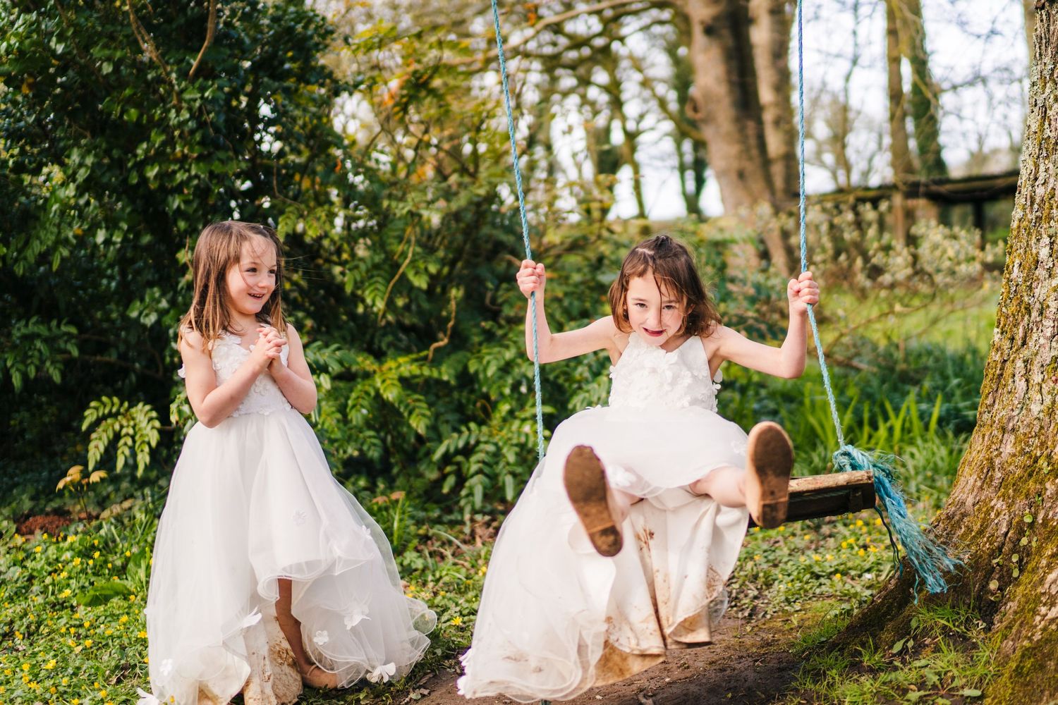 Two young children in white dresses playing outdoors in a lush garden setting with a wooden swing. family PHOTOGRAPHER Dublin