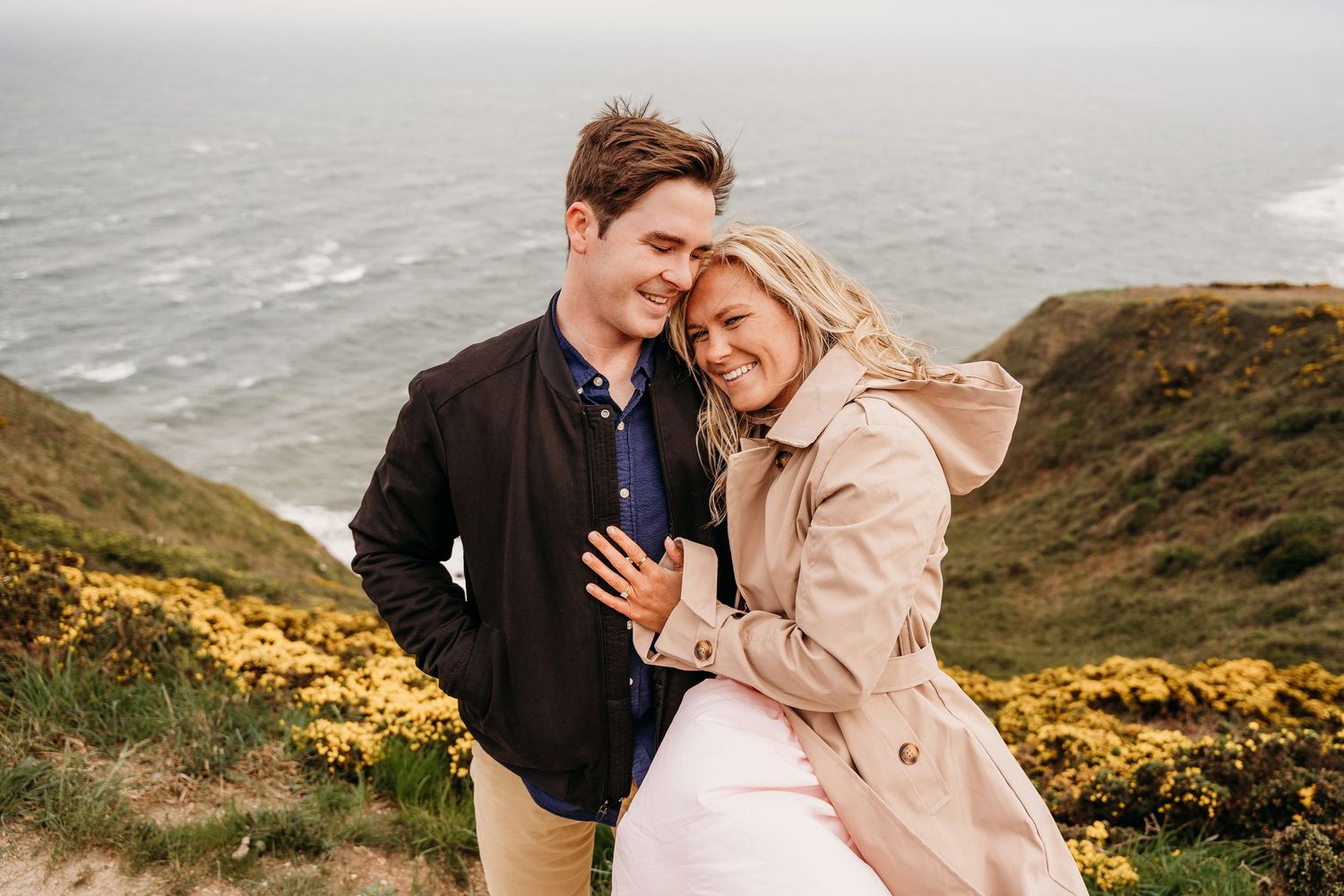 A couple sharing an intimate moment on a rugged coastal cliff overlooking the ocean on a cloudy day. PHOTOGRAPHER Dublin ireland