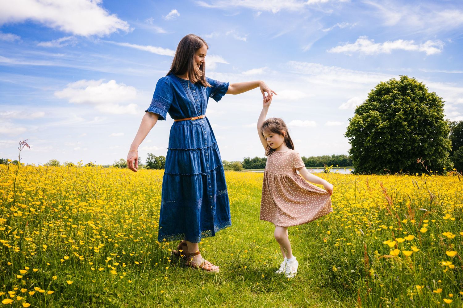Two people in dresses dance together in a vibrant yellow wildflower meadow on a sunny summer day. family PHOTOGRAPHER Dublin