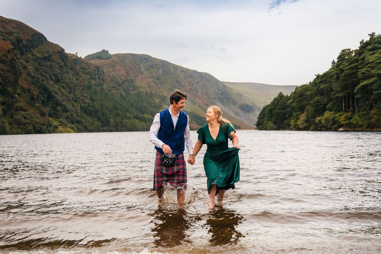 Couple walking together in shallow water along a scenic mountain lake with misty hills in the background.Family photogrAPHER DUBLIN