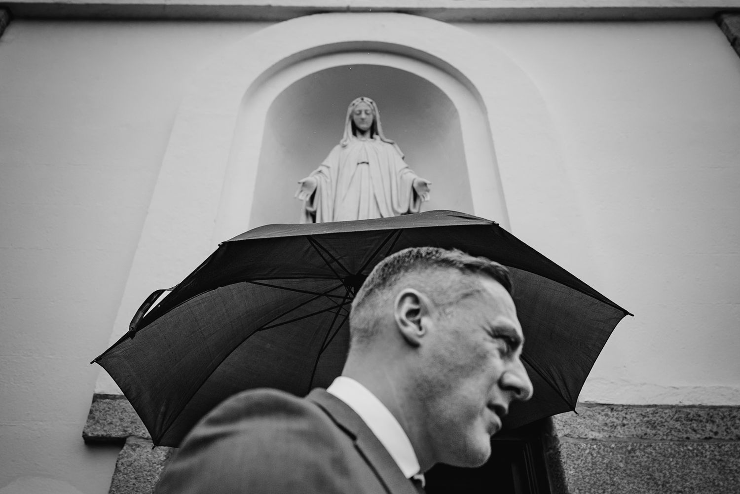 A person holding an umbrella stands beneath a religious statue mounted in an arched alcove in black and white. The Dean Dublin Wedding Photo