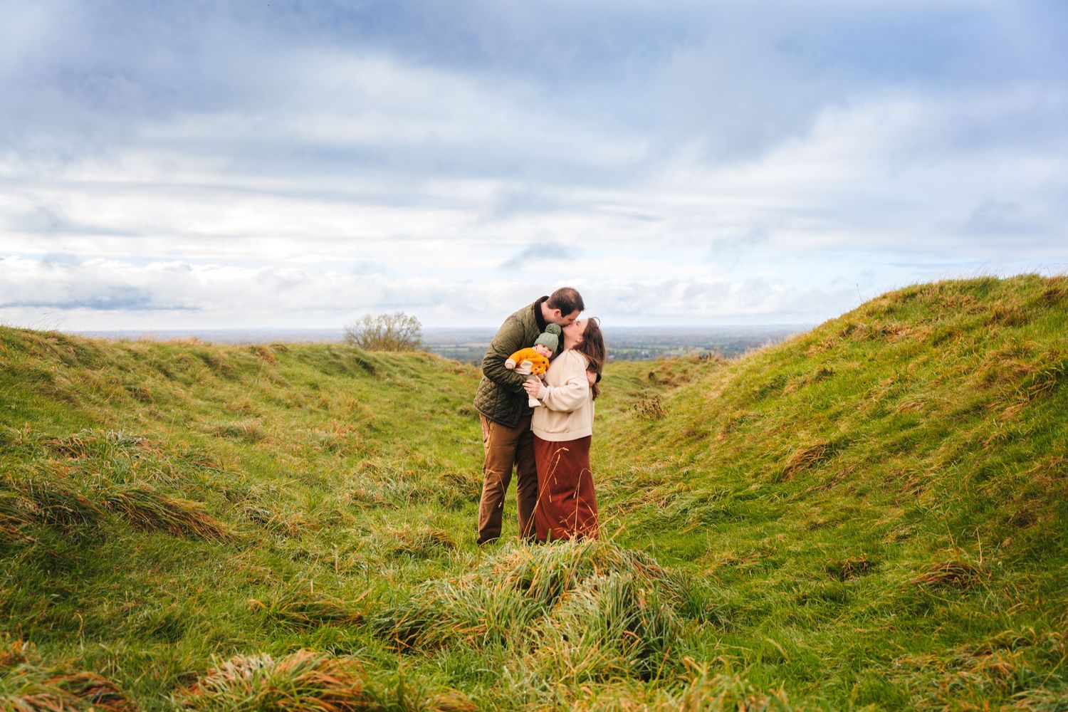 Couple embraces in scenic rolling green hills with dramatic cloudy sky in background. Family photographer Dublin