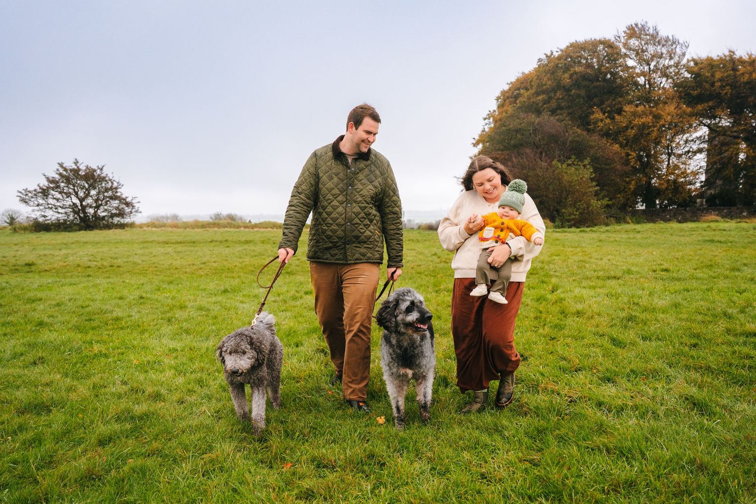 A couple and their two dogs walk together through a green hillside meadow on an autumn afternoon. Family photographer Dublin