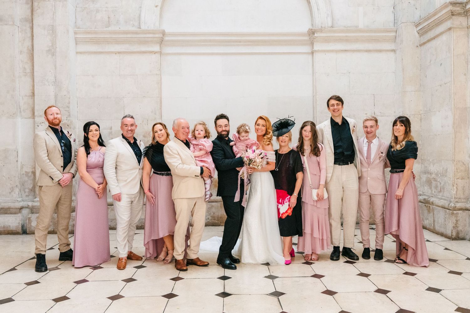 Wedding party members in pink dresses and dark suits pose together against a light stone wall backdrop.