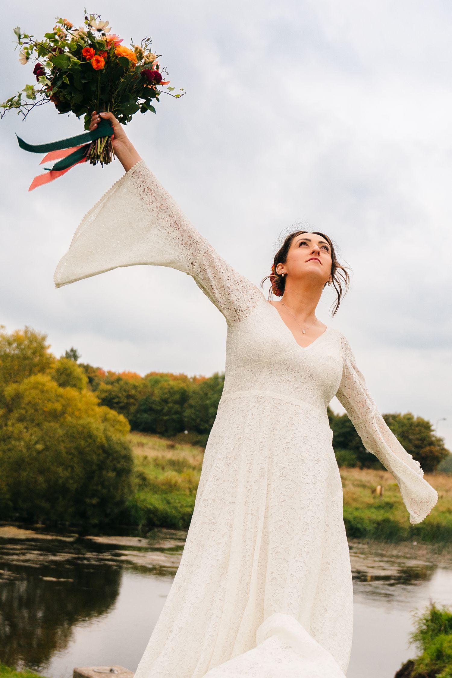 White-dressed figure dances joyfully by a riverbank holding flowers against an autumn backdrop. Wedding Photography Trim Castle Ireland