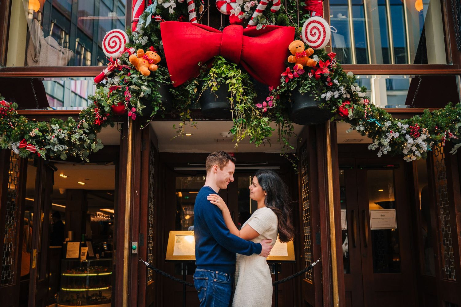 A couple embraces in front of storefronts decorated with festive red and floral holiday decorations. Surprise proposal dublin photography