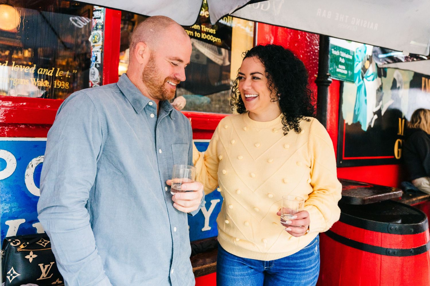 A couple shares a laugh while holding drinks at a casual outdoor establishment with red decor. Proposal Dublin Photographer