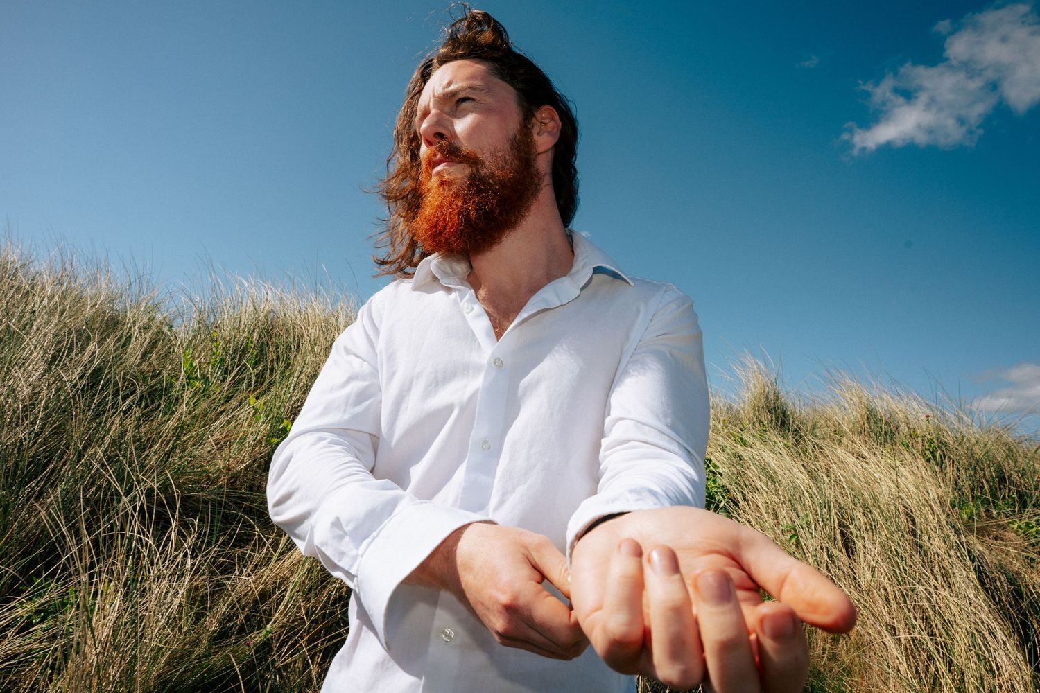 Musician with a guitar sitting in tall grass against a blue sky with white clouds. PHOTOGRAPHER Dublin