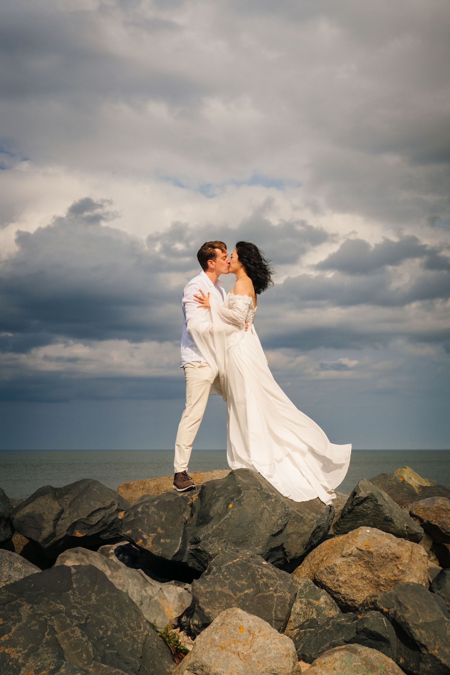 A couple embraces on rocky coastal terrain with dramatic cloudy skies and ocean backdrop at sunset.