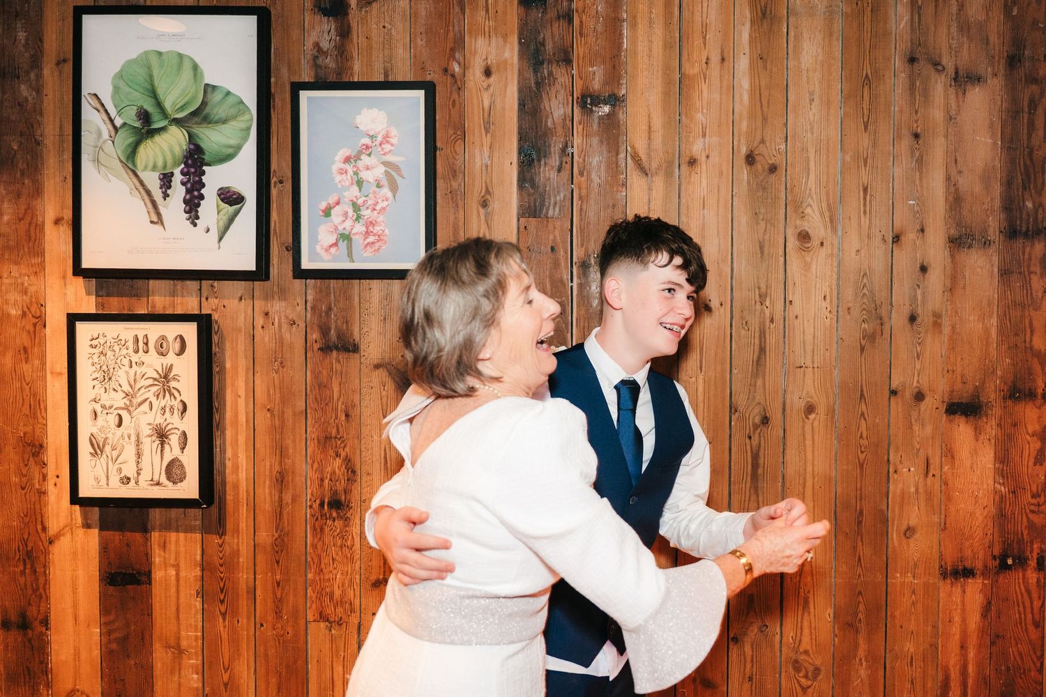 A couple dances together in front of a rustic wooden wall decorated with framed botanical prints. The Dean Dublin Wedding Photographer