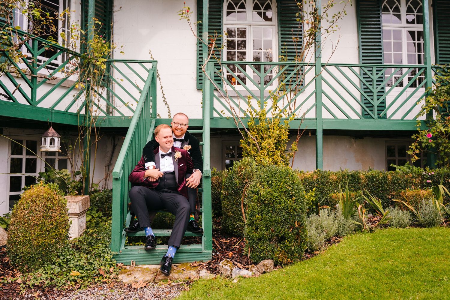 A couple sits together on the steps of a white house with green shutters. LGBTQ Ashley Park Wedding Photographer