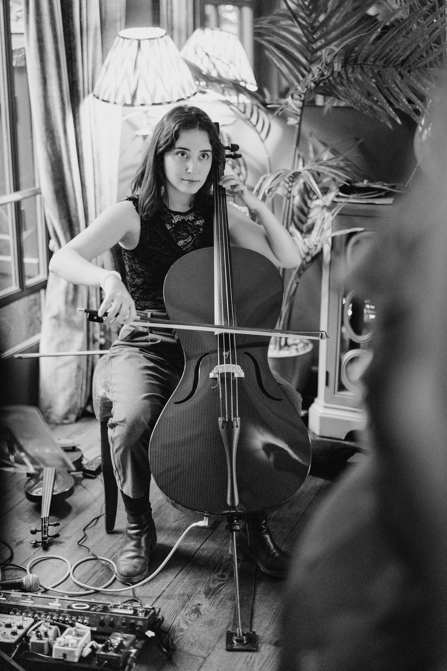 Black and white image of a musician playing cello with guitar pedals on wooden floor in a room with decorative lamps. Marco Pierre Dublin We