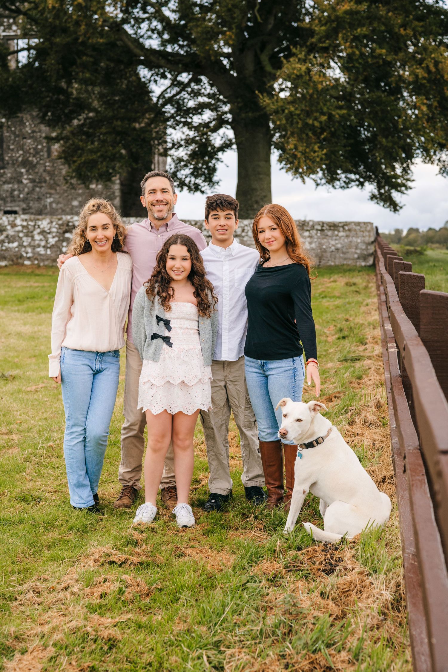 A group poses casually together outdoors against a rustic wooden fence on grassy terrain. BEctive abbey family photographer dublin