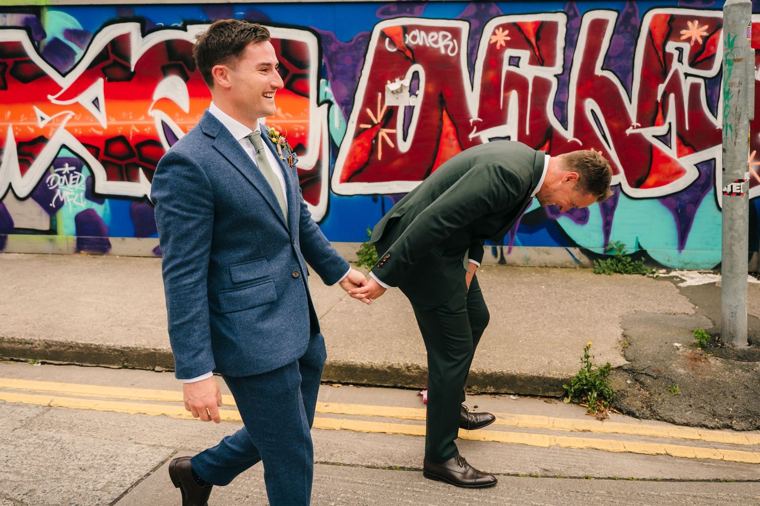 Two people in suits posing playfully in front of colorful street art graffiti. Same sex wedding PHOTOGRAPHER Dublin