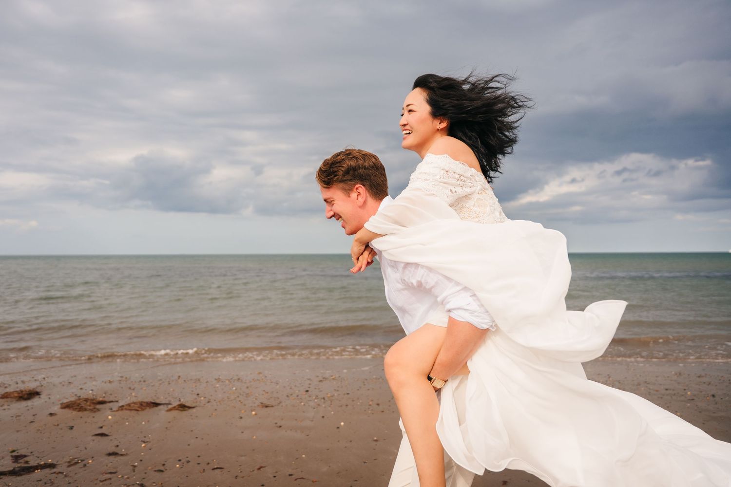 A dreamy coastal wedding photoshoot shows a couple standing by the ocean as a white dress billows in the wind.