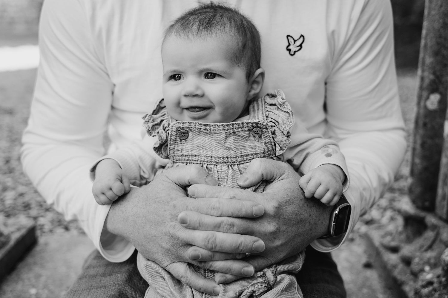 Black and white portrait of a smiling baby wearing overalls being held by supportive hands. Family PHOTOGRAPHER Dublin