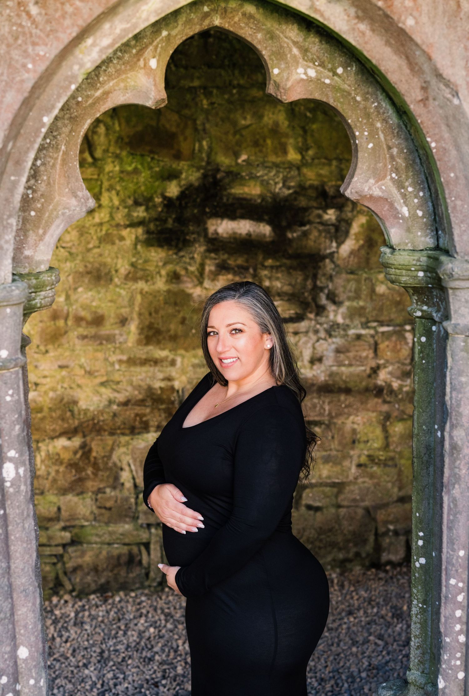 Maternity photo of woman in black dress posing under ornate stone archway. Family PHOTOGRAPHER Dublin