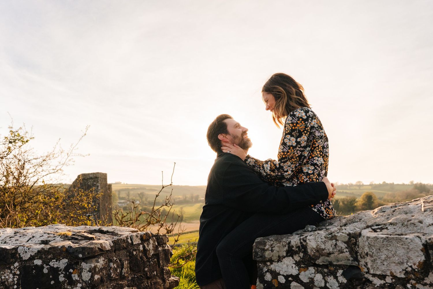 Couple sharing an intimate moment on stone wall with scenic countryside view during sunset. Photographer Ireland dublin