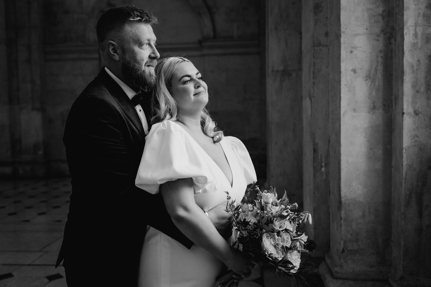 A romantic black and white wedding portrait of a couple embracing near stone columns with the bride holding a floral bouquet. City Hall Dubl