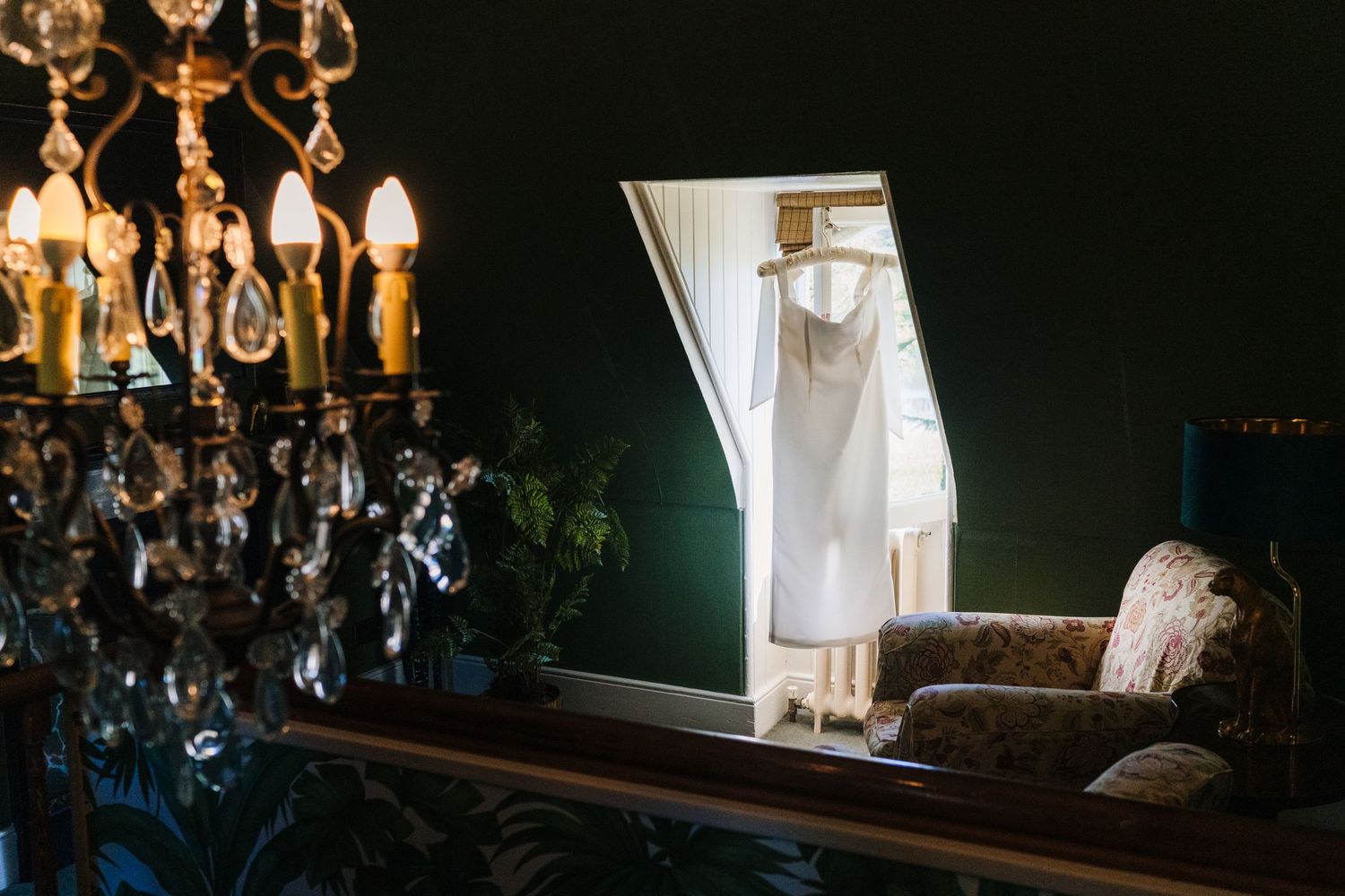 An elegant white wedding dress hangs illuminated in a doorway with a crystal chandelier in the foreground. Castle Leslie Wedding Photographe