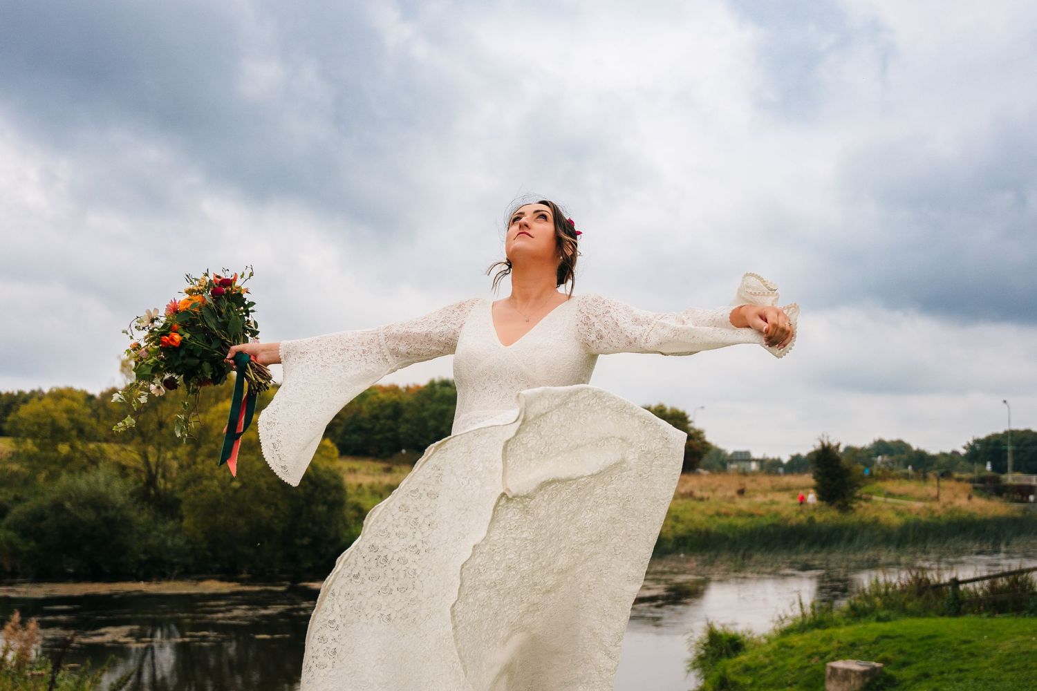 Bride in lace dress poses playfully by a river on a cloudy day. Wedding Photography Trim Castle Ireland