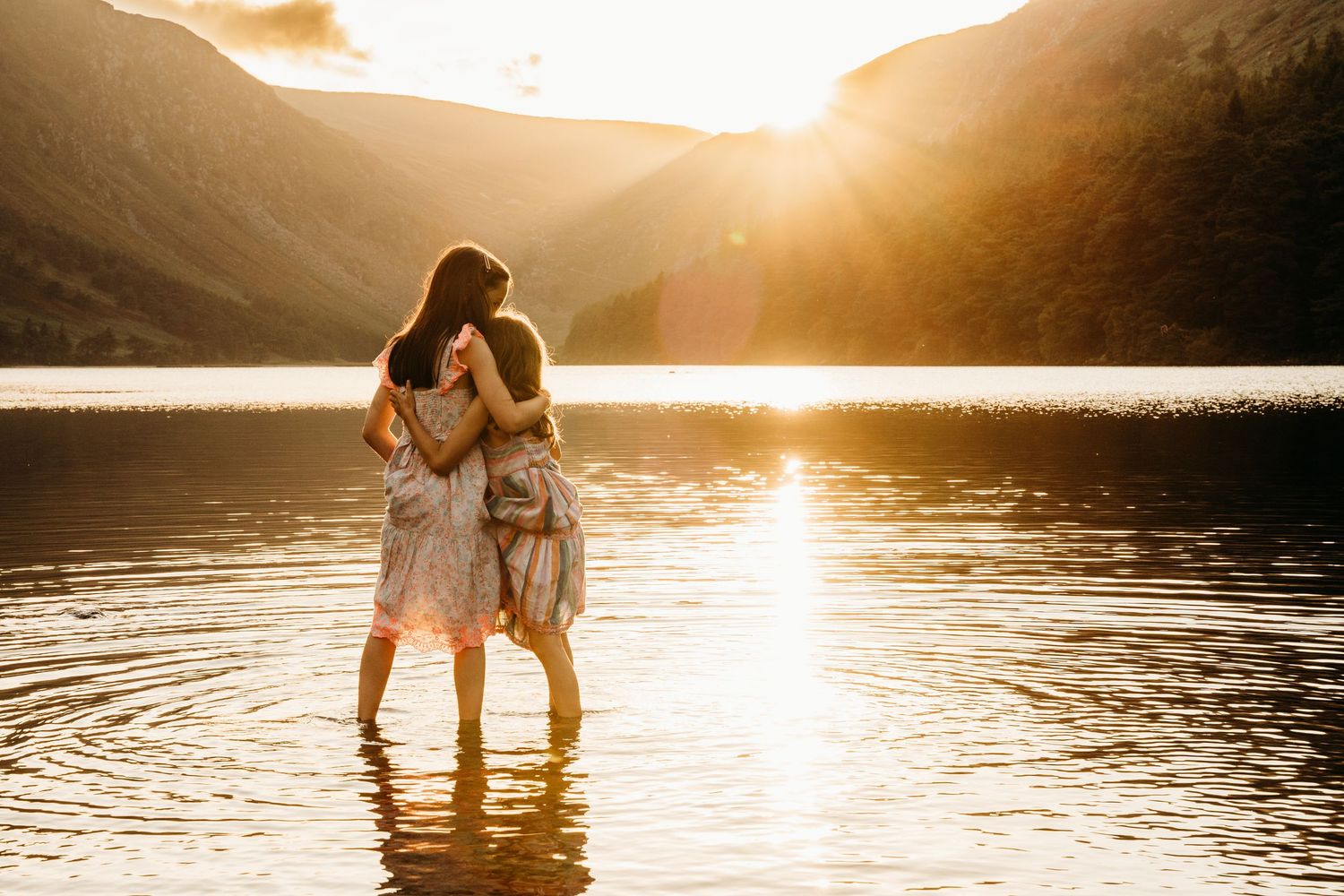 A couple stands together in silhouette against a golden sunset reflecting on calm lake waters. Glendalough family photographer