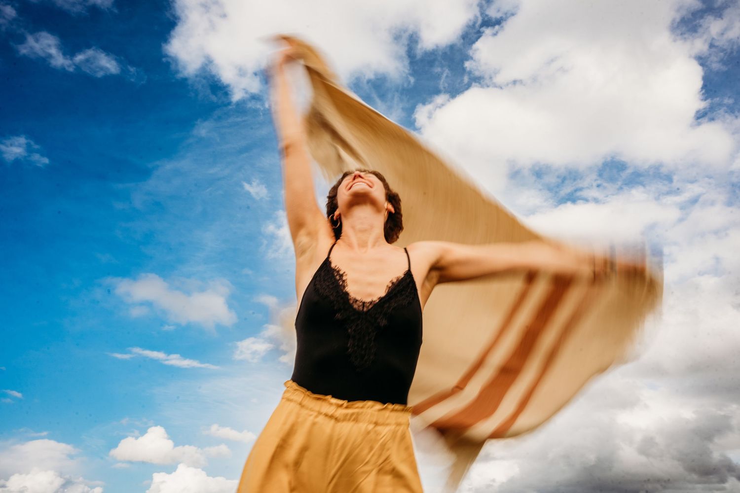 Figure in a black tank top and flowing yellow skirt dances against blue sky with white clouds. PHOTOGRAPHER Dublin ireland