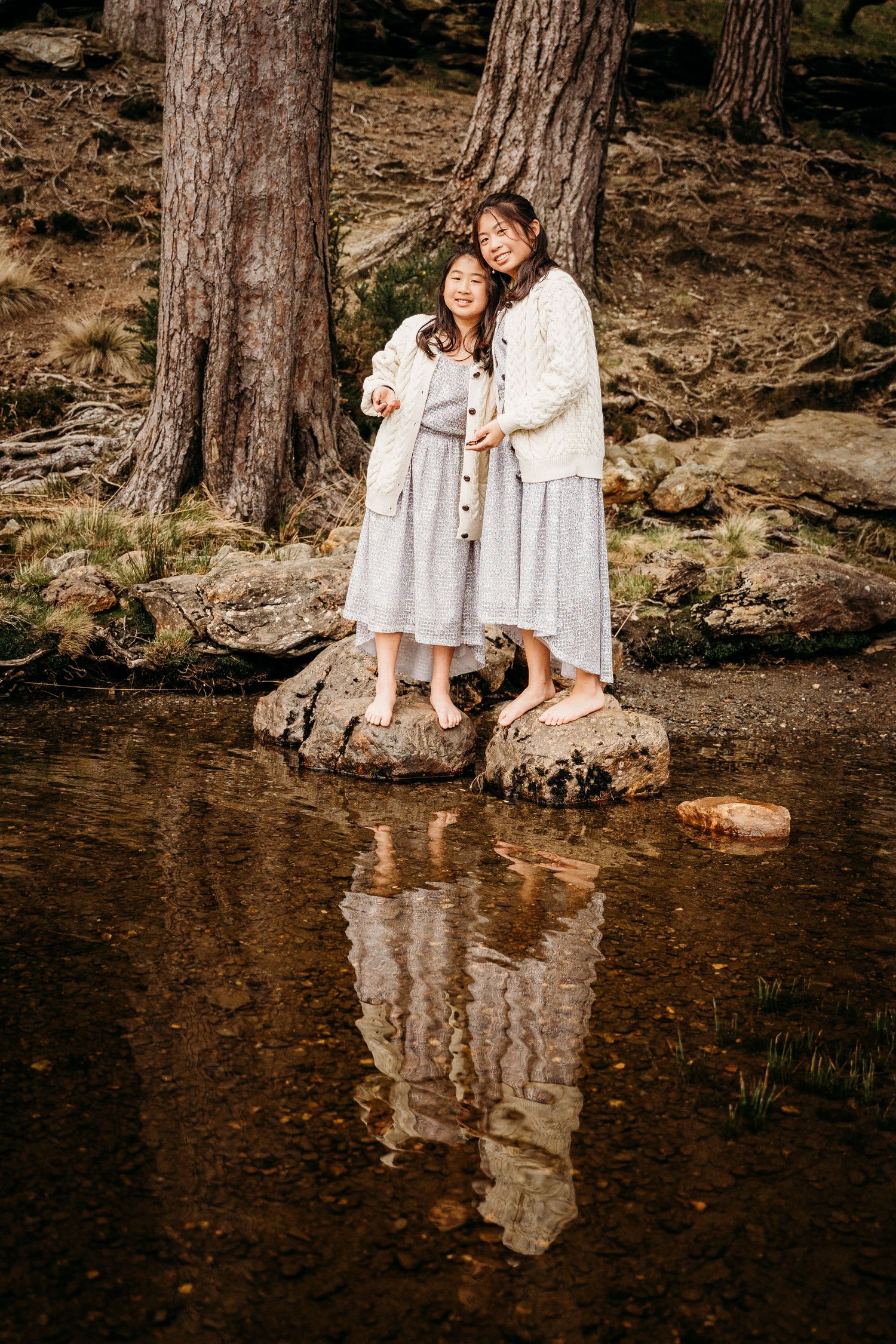 Two people in white dresses stand on rocks by a reflecting pond in a forest setting with tall pine trees. family PHOTOGRAPHER Dublin