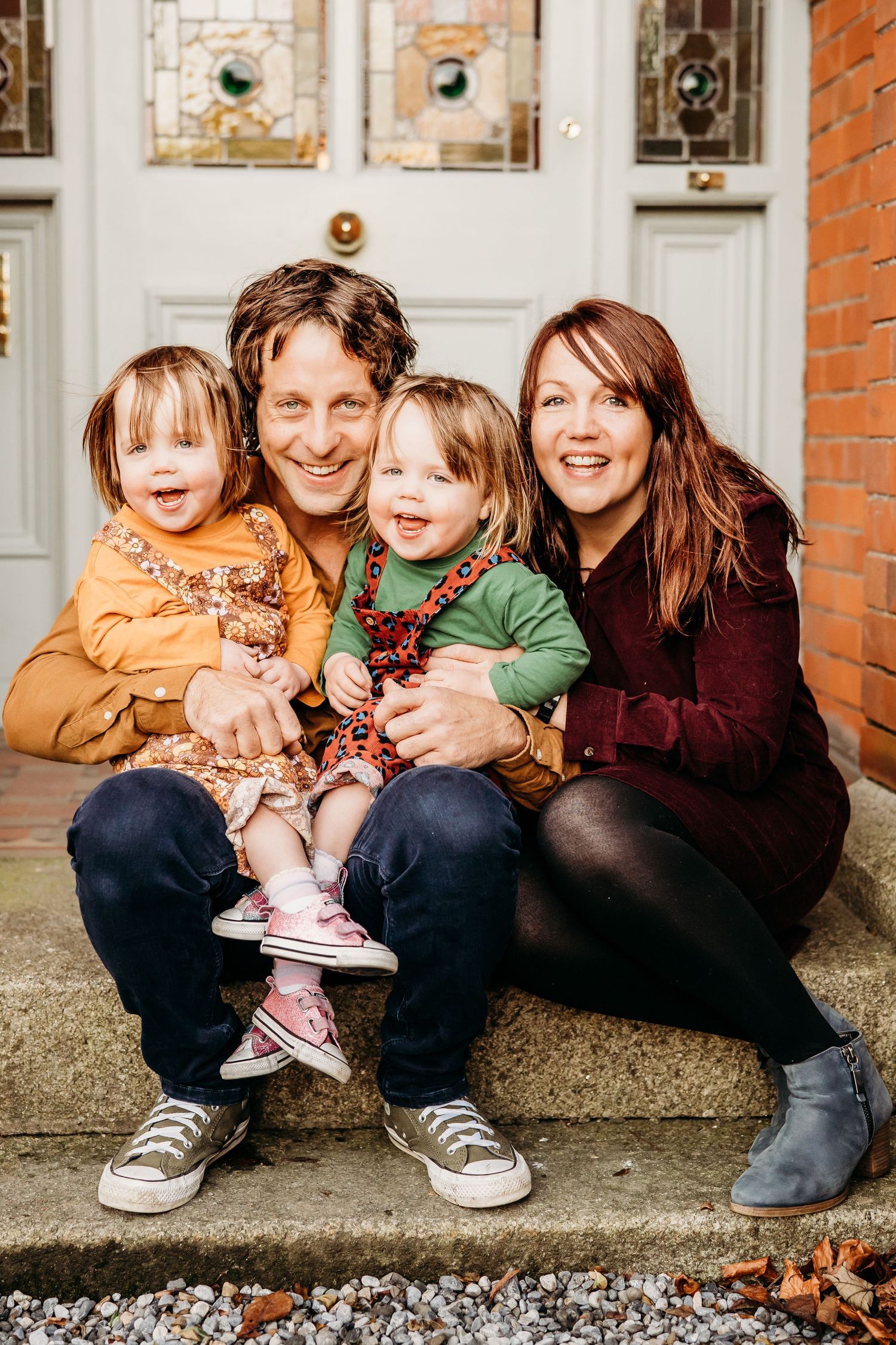 A family sits together on stone steps in front of a white door during autumn, all wearing casual clothing and smiling. family PHOTOGRAPHER D