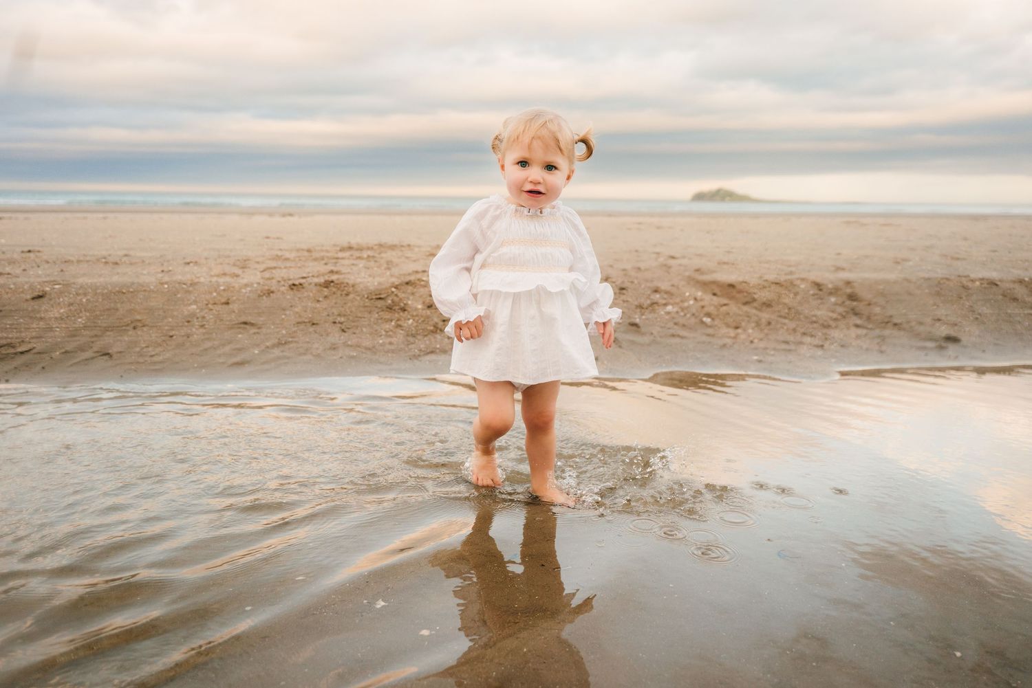 Small figure in white dress with rain boots joyfully standing in shallow tide waters on cloudy beach. family PHOTOGRAPHER Dublin