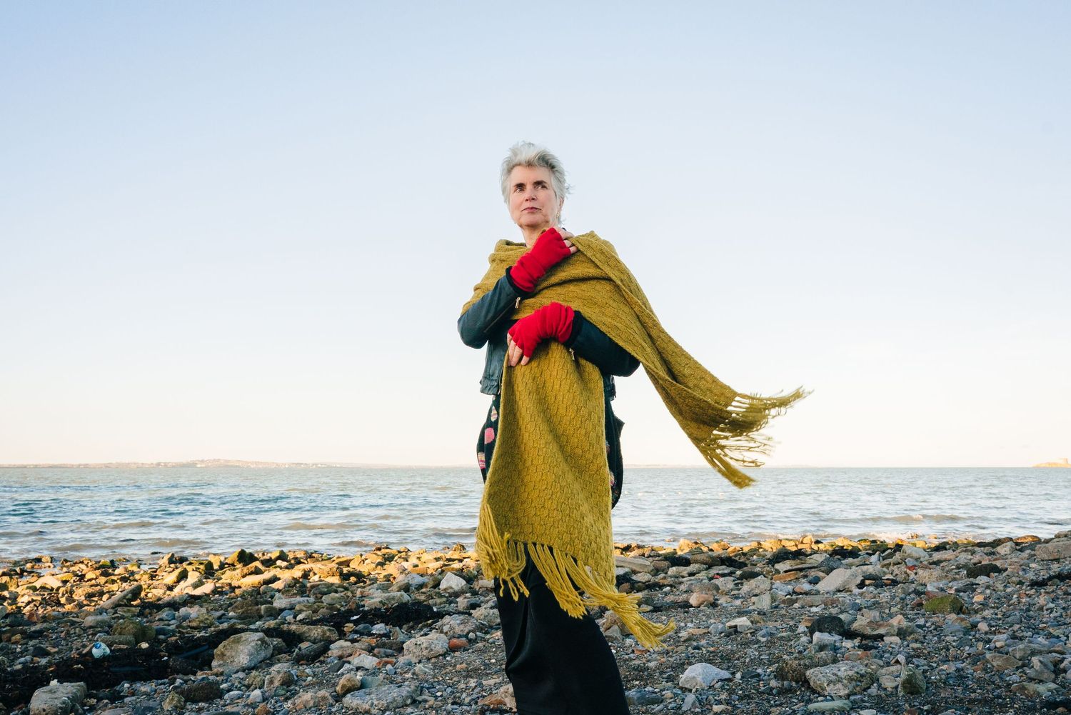 Person in olive green coat and red scarf walks along rocky beach shoreline. professional headshots branding photographer dublin ireland