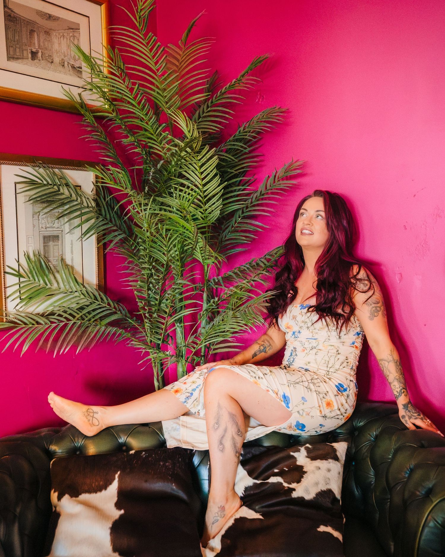 Woman in floral dress lounges playfully on dark couch with pink wall and palm plant backdrop. LGBTQ Ashley Park Wedding Photographer